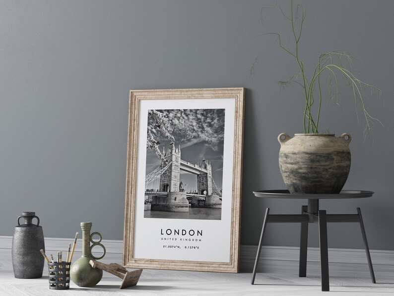 May include: A framed black and white print of the Tower Bridge in London, United Kingdom, with the city's name and coordinates. The print has a light wood frame and is leaning against a gray wall. A decorative vase and other objects are in the foreground.