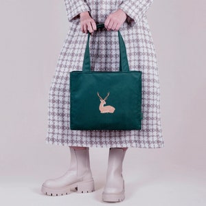 Könnte beinhalten: Eine grüne Einkaufstasche mit einem Hirsch-Design. Die Tasche hat zwei Henkel und ist aus Canvas gefertigt.