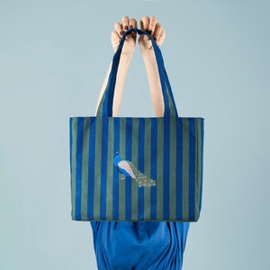 May include: A blue and green striped tote bag with a peacock design. The bag has two handles and is being held by a person.