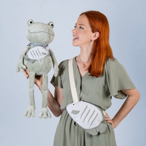 May include: A woman wearing a green dress holds a plush frog toy with a fish design. She is also wearing a crossbody bag with a fish design.