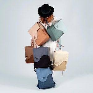 May include: A woman wearing a black hat holds a stack of six different colored canvas backpacks. The backpacks have various embroidered designs, including a bird, a whale, a deer, and a plant.