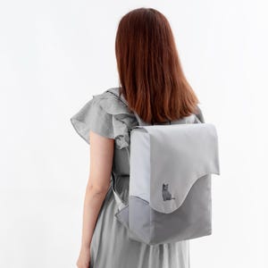 Puede incluir: Una mochila gris con cierre de solapa y diseño bicolor. La solapa superior es gris claro y la sección inferior es gris más oscuro. Un pequeño gato bordado está en la parte delantera. La mochila tiene correas ajustables.