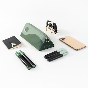 May include: A green and white triangular pencil case with the word "VIFA" printed on it. The case is open and there are two black and green markers inside. There is a small toy cow, a notebook with a picture of a person on it, and a gold phone on the white surface.