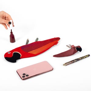 Puede incluir: Un estuche rojo con forma de loro con rayas amarillas y azules, un llavero a juego y un teléfono inteligente rosa se exhiben sobre una superficie blanca. También hay un frasco de esmalte de uñas rojo y un bolígrafo.