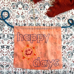 May include: A peach-colored fabric banner with the words "happy days" embroidered in blue thread. A yellow sun with a smiling face is embroidered below the text.