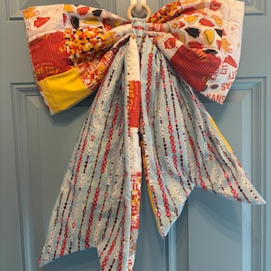 May include: A large fabric bow featuring a patchwork design with red, yellow, and white Kansas City football team patterns. The long, dangling tails are light blue with repeating text including "Red Kingdom Era" and "What a Catch" in red and black.