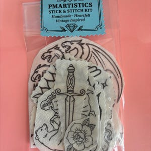 May include: A clear plastic bag containing a stick and stitch kit. The kit includes several white fabric patches with black line drawings of a dragon, sword, rose, and other vintage-inspired designs. The kit is labeled "PMARTISTICS STICK & STITCH KIT".