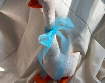 Könnte beinhalten: Ein ausgestopftes Gänse-Stofftier aus weißem Stoff mit einem gesprenkelten Muster in Orange- und Brauntönen. Die Gans hat einen leuchtend orangefarbenen Schnabel und orangefarbene Füße sowie ein transparentes, hellblaues Band, das um den langen Hals gebunden ist.