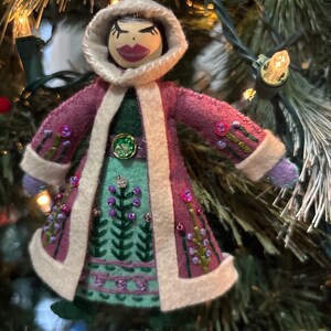 May include: A felt doll ornament featuring a wooden head with painted facial features and a hood. The doll wears a plum-colored coat with cream-colored trim, a green skirt with embroidered floral patterns, and small beads, hanging by a thin string.