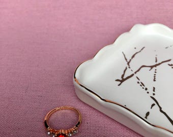 May include: A white ceramic ring dish with a gold rim and a gold tree design. A red and clear crystal ring is sitting on the dish.