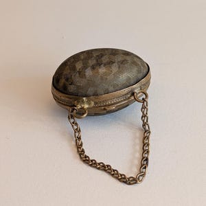 May include: An antique, oval trinket box with a textured, geometric pattern on the lid. The box is made of metal with a tarnished, bronze finish. A delicate chain is attached for carrying.