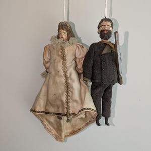 May include: Two vintage hanging ornaments. One is a woman in a pink dress with a lace collar and gold trim. The other is a man in a dark suit holding a wooden rifle. Both have painted faces and are suspended by white strings.