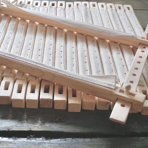 May include: A rectangular embroidery frame constructed from light-colored wood, featuring adjustable side bars with multiple pre-drilled holes and wooden pegs. The frame includes strips of off-white fabric with small metal tacks for securing fabric.
