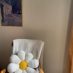 K&ouml;nnte beinhalten: Ein gro&szlig;er, handgefertigter G&auml;nsebl&uuml;mchen-Kissen mit wei&szlig;en Bl&uuml;tenbl&auml;ttern und einem gelben Zentrum liegt auf einem Holzstuhl. Der Stuhl ist mit einer wei&szlig;en Decke bedeckt. Ein gerahmtes Gem&auml;lde h&auml;ngt an der Wand.