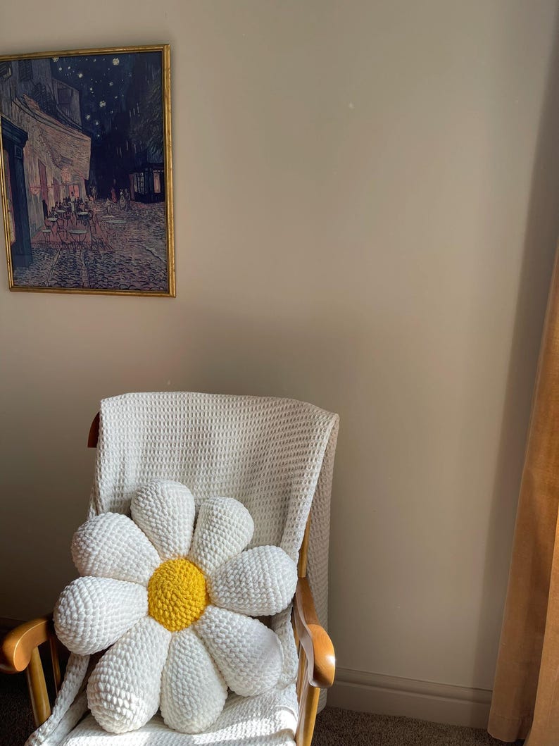 K&ouml;nnte beinhalten: Ein gro&szlig;er, handgefertigter G&auml;nsebl&uuml;mchen-Kissen mit wei&szlig;en Bl&uuml;tenbl&auml;ttern und einem gelben Zentrum liegt auf einem Holzstuhl. Der Stuhl ist mit einer wei&szlig;en Decke bedeckt. Ein gerahmtes Gem&auml;lde h&auml;ngt an der Wand.