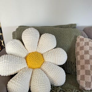 K&ouml;nnte beinhalten: Ein gro&szlig;es, wei&szlig;es kissenf&ouml;rmiges G&auml;nsebl&uuml;mchen mit gelber Mitte. Das Kissen besteht aus einem weichen, strukturierten Material und liegt auf einem grauen Sofa. Eine gr&uuml;ne Decke mit Fransen und ein kariertes Kissen sind ebenfalls zu sehen.