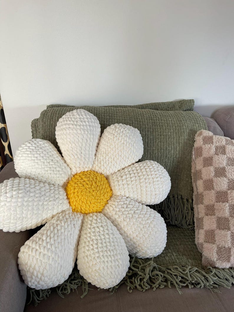 K&ouml;nnte beinhalten: Ein gro&szlig;es, wei&szlig;es kissenf&ouml;rmiges G&auml;nsebl&uuml;mchen mit gelber Mitte. Das Kissen besteht aus einem weichen, strukturierten Material und liegt auf einem grauen Sofa. Eine gr&uuml;ne Decke mit Fransen und ein kariertes Kissen sind ebenfalls zu sehen.