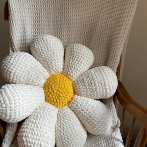 K&ouml;nnte beinhalten: Ein gro&szlig;es, handgefertigtes G&auml;nsebl&uuml;mchen-Kissen mit wei&szlig;en Bl&uuml;tenbl&auml;ttern und einem gelben Zentrum. Das Kissen besteht aus einem weichen, strukturierten Material und liegt auf einem Holzstuhl mit einer wei&szlig;en Decke. Es ist ein dekorativer Artikel.