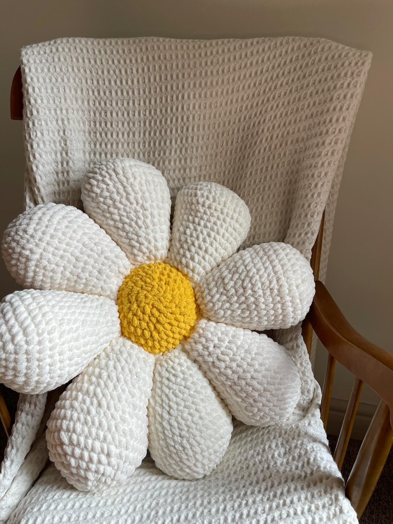 K&ouml;nnte beinhalten: Ein gro&szlig;es, handgefertigtes G&auml;nsebl&uuml;mchen-Kissen mit wei&szlig;en Bl&uuml;tenbl&auml;ttern und einem gelben Zentrum. Das Kissen besteht aus einem weichen, strukturierten Material und liegt auf einem Holzstuhl mit einer wei&szlig;en Decke. Es ist ein dekorativer Artikel.