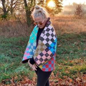 May include: A colorful patchwork quilt wrap with a variety of patterned squares in shades of blue, pink, and brown. The wrap is worn over a black top and leggings, paired with black fringed boots. The background features a natural outdoor setting.