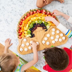 May include: A rainbow fruit platter with strawberries, oranges, pineapple, kiwi, grapes, and blueberries. The platter is shaped like a rainbow and has two cloud-shaped sections with white meringue cookies.