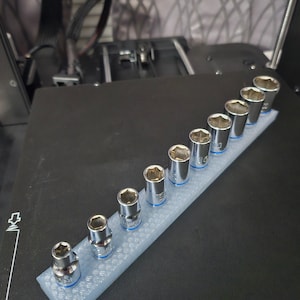 May include: A white plastic organiser with a grid pattern holds a set of nine silver socket wrenches. The wrenches are arranged in order of size, with the smallest wrench on the left and the largest wrench on the right.