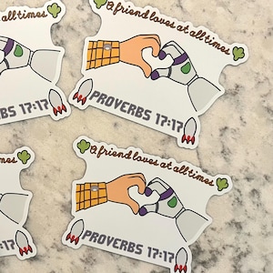 May include: White stickers with a cartoon hand and robot hand forming a heart shape. The text "A friend loves at all times" and "Proverbs 17:17" are included. The stickers have a rocket ship and green clover design.