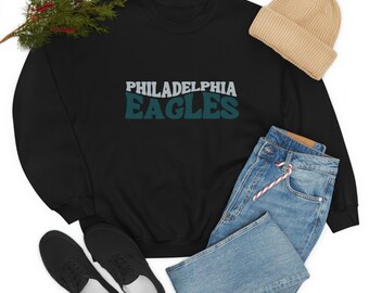 May include: Black sweatshirt with a green and white "Philadelphia Eagles" logo. The sweatshirt is laid out on a white background with a pair of black shoes, blue jeans, and a beige beanie.