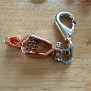 May include: A copper-colored metal spring clamp attached to a silver-toned swivel snap hook. The clamp features a visible internal metal coil spring and a serrated jaw design, connected to the hook by a small metal shackle and pin assembly.