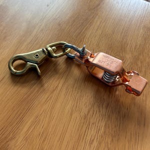 Op de afbeelding: Een metalen klem met een veer en een goudkleurige sluiting. De klem is koperkleurig en heeft de tekst "MUELLER 75-AMP". De sluiting is met een zilverkleurige ring aan de klem bevestigd.