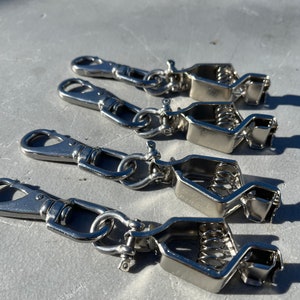 May include: Four silver metal clips with spring-loaded jaws and a swivel hook. The clips are designed for attaching items to a surface.