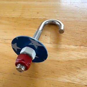 May include: A metal J-hook bolt featuring a blue circular washer decorated with white stars. The assembly includes a white hexagonal nut and a red nylon-insert lock nut threaded onto the bolt shaft, creating a secure and functional hardware component.