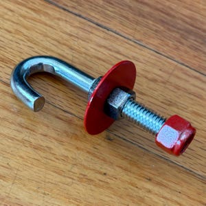 May include: A silver metal J-hook bolt featuring a circular red washer, a silver hexagonal nut, and a red cap nut at the threaded end. The hardware component has a smooth metallic finish on the hook portion and a threaded shaft for secure fastening.