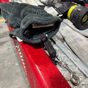 Puede incluir: Un guante de trabajo de cuero negro con un clip negro y un mosquetón plateado. El guante descansa sobre una superficie roja con un patrón de placa de diamante. El guante tiene una etiqueta en el interior con texto.