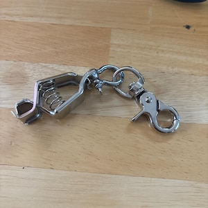 Puede incluir: Un mosquetón de llavero de metal plateado brillante con una abrazadera con resorte y un cierre giratorio. El clip está conectado a un pequeño anillo. Un tornillo pequeño y unos alicates están en el fondo.