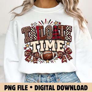 Może przedstawiać: Biała bluza z okrągłym dekoltem z nadrukiem graficznym z napisem TAILGATE TIME w blokowych literach. Litery mają wzory w panterkę, kratę i szachownicę. Pod napisem znajduje się brązowa piłka do futbolu amerykańskiego oraz dwie kokardy.