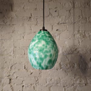 Può includere: Una lampada a sospensione in vetro verde con macchie bianche. La luce è appesa a un cavo marrone.