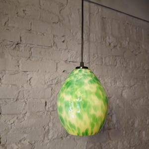 Può includere: Una lampada a sospensione in vetro verde e giallo con un cavo nero appeso a un muro di mattoni bianchi.