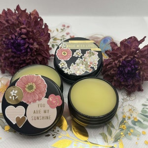 Puede incluir: Tres pequeños botes redondos con bálsamo labial. Los botes son negros con detalles dorados. Un bote tiene un diseño floral y el texto "Made with Love". Otro bote tiene un diseño floral y el texto "You are my sunshine". El tercer bote contiene bálsamo labial amarillo.