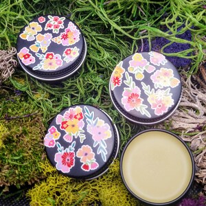 May include: Three small round tins with black lids and floral designs. The tins contain a yellow balm.