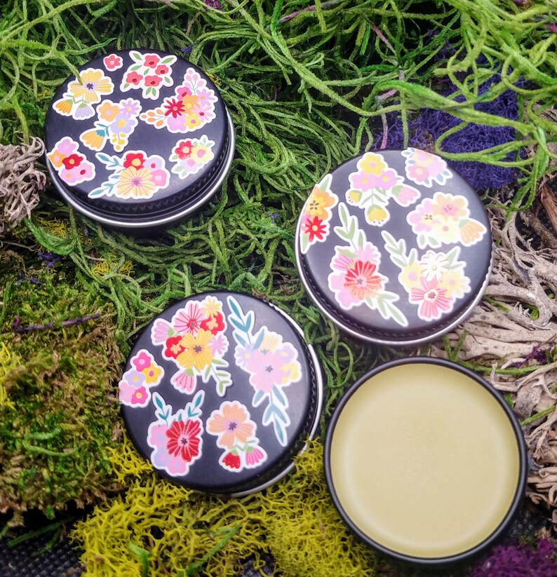 May include: Three small round tins with black lids and floral designs. The tins contain a yellow balm.