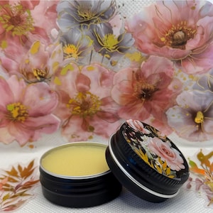 May include: A small, round tin container filled with a yellow balm. The tin has a black lid with a floral design. The tin is sitting on a white surface with a floral pattern.