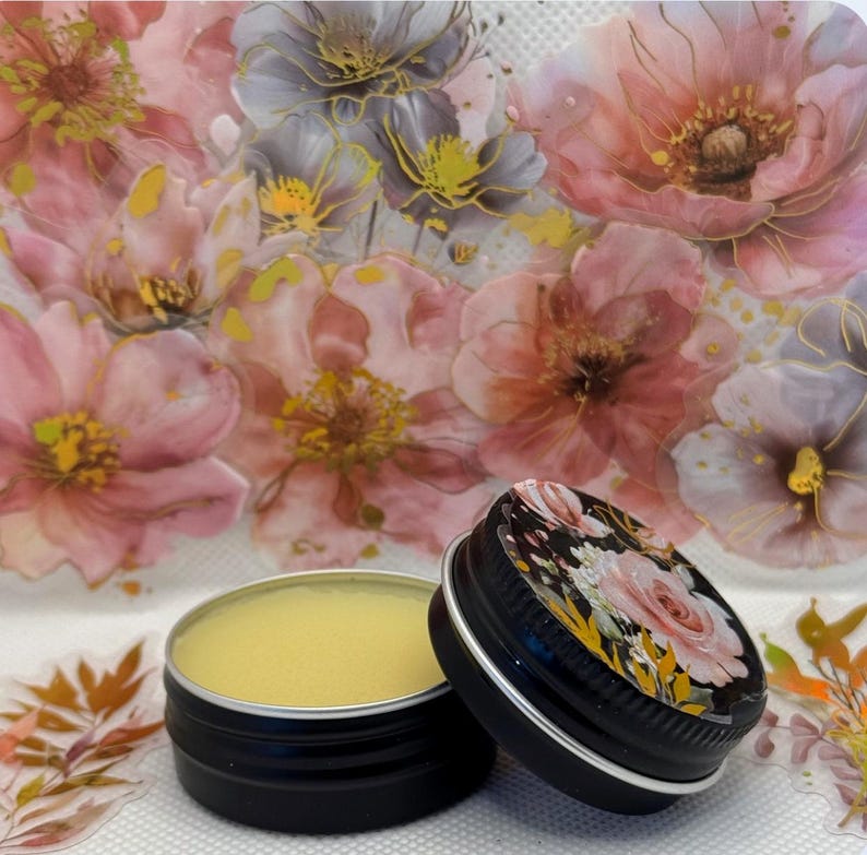 May include: A small, round tin container filled with a yellow balm. The tin has a black lid with a floral design. The tin is sitting on a white surface with a floral pattern.