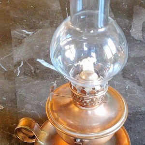 Könnte beinhalten: Eine Kupfer-Öllampe mit einem Glas-Schornstein und einem Griff. Die Lampe hat ein dekoratives Design auf der Basis und einen Docht im Schornstein.