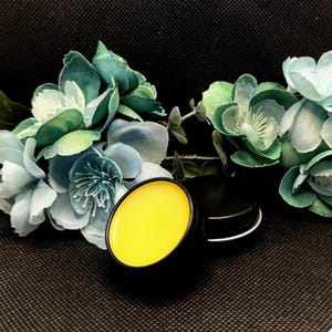 Może przedstawiać: Mały, okrągły pojemnik z blachy z żółtym balsamem do ust w środku. Pojemnik jest otoczony niebieskimi i zielonymi sztucznymi kwiatami.