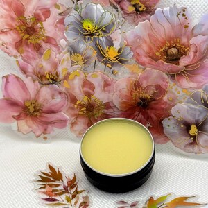 May include: A small round tin container with a lid, filled with a yellow balm. The container is sitting on a white surface with a floral design.