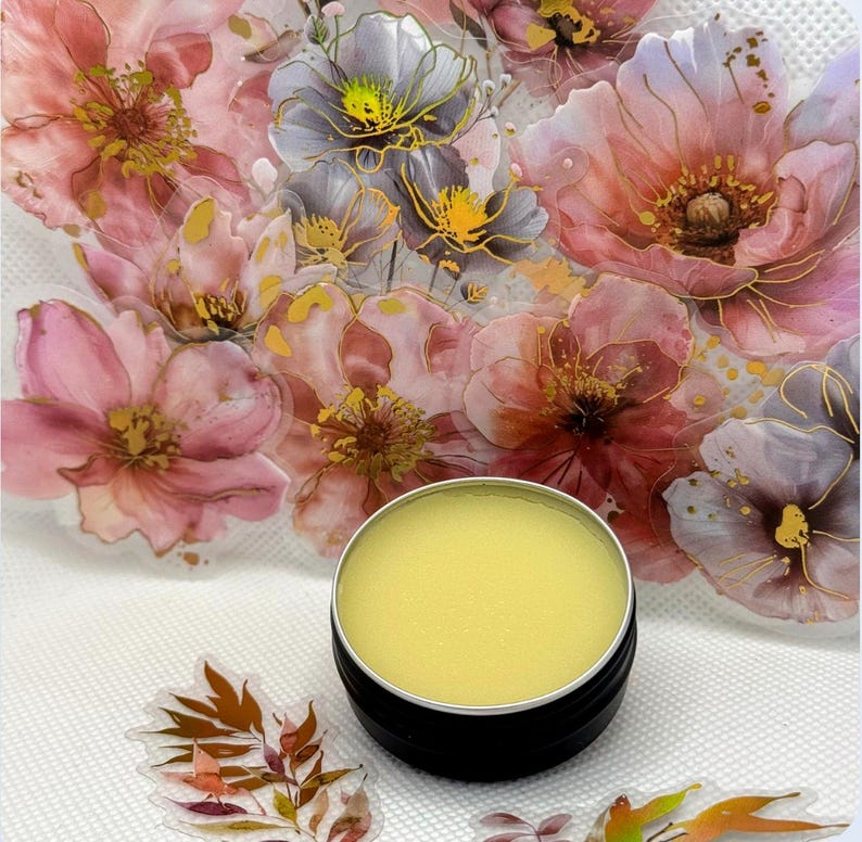 May include: A small round tin container with a lid, filled with a yellow balm. The container is sitting on a white surface with a floral design.