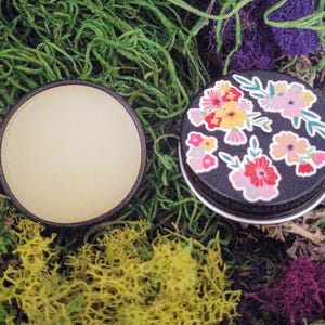 May include: A small, round tin with a black lid and a floral design. The lid is decorated with colorful flowers and leaves. The tin is open and the inside is empty.