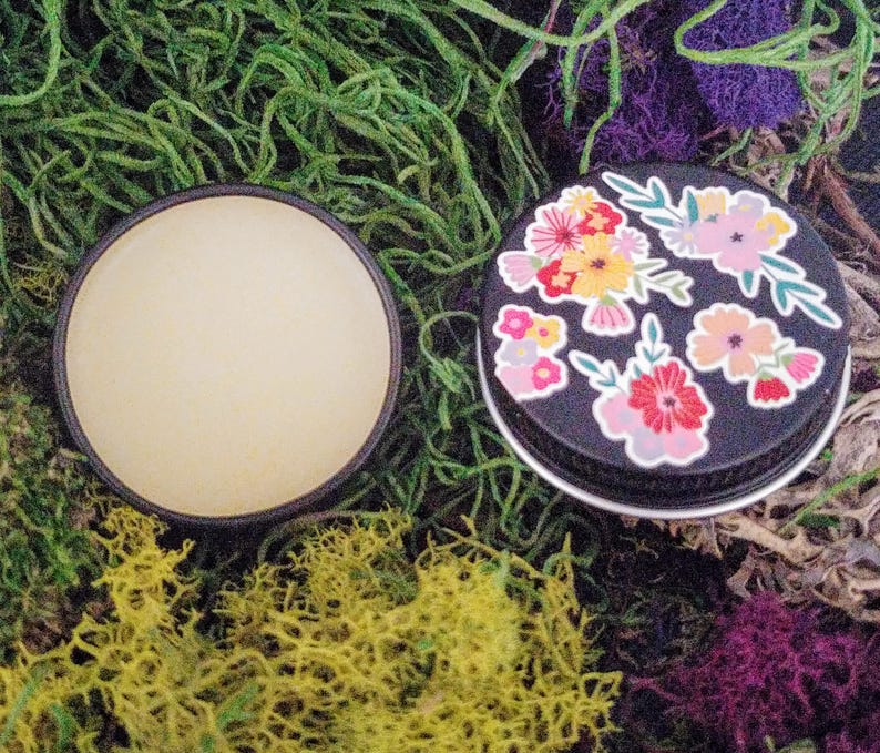 May include: A small, round tin with a black lid and a floral design. The lid is decorated with colorful flowers and leaves. The tin is open and the inside is empty.