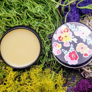 May include: A small round tin with a black lid and a floral design. The lid is open and the tin contains a yellow balm.